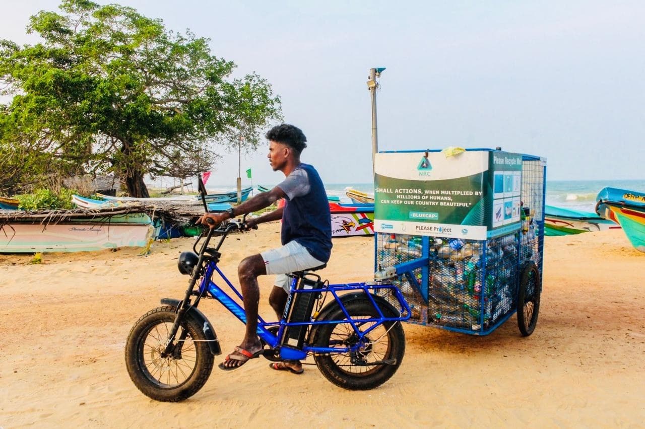 An NRC collector riding an electric cargo bicycle with a BLUECAP collection trailer full of bottles along the beach.